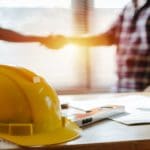 Construction work hat sitting on table in front of two AV subcontractors shaking hands.