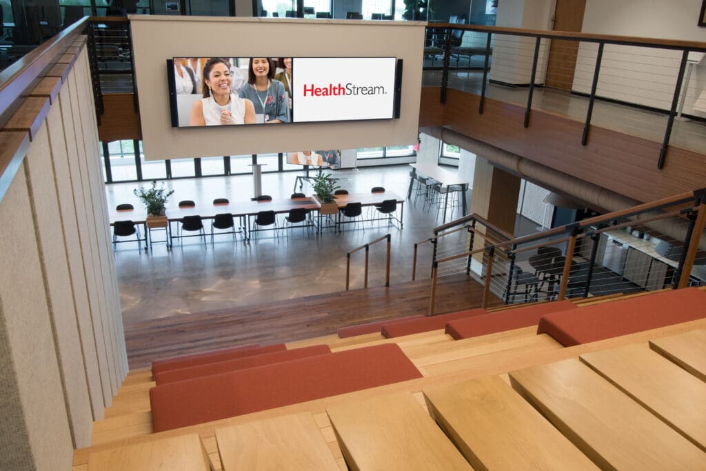 A stairway that is also fashioned a seating arrangement. The stairs lead down to a platform with a conference table. Display screens are placed on the walls opposite the stairway.