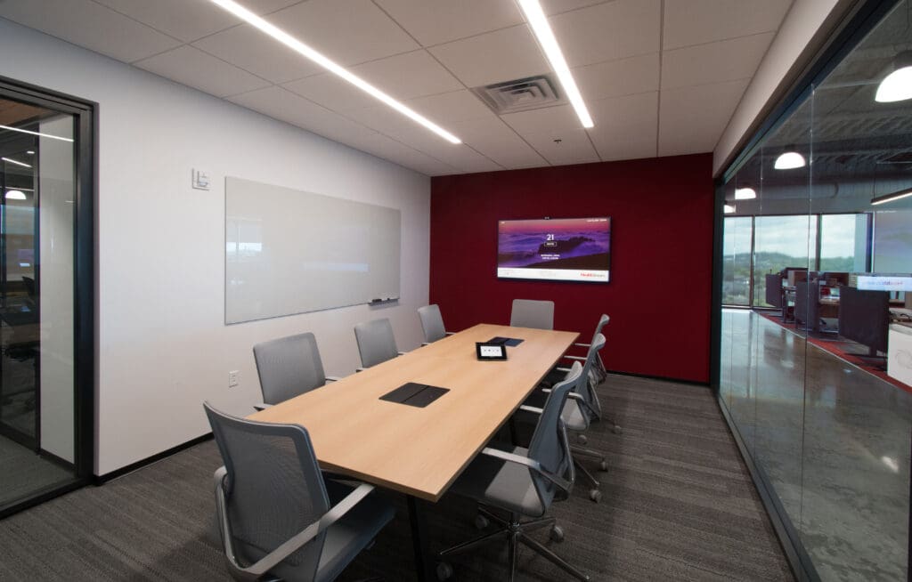 Office room with a glass wall. The room also features another red wall with a large display screen. In the center, a long table has chairs on either side.