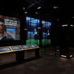 A dark room with multiple large screen showing rugby players. A smaller panel of touchscreens is placed in the middle of the room.