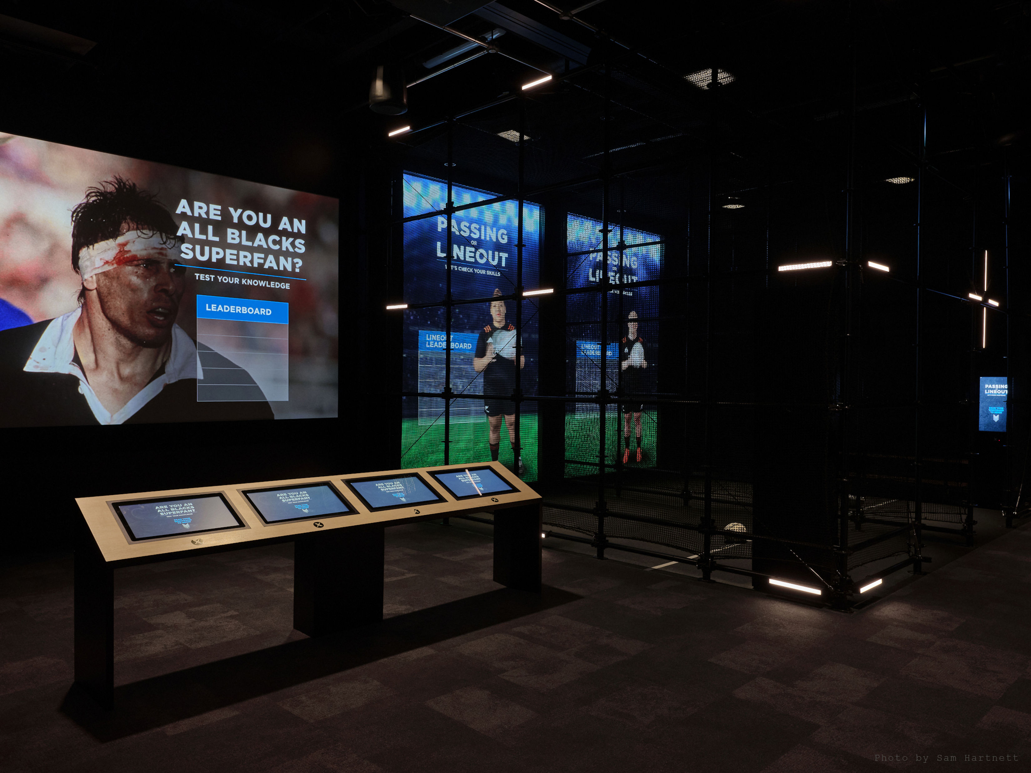 A dark room with multiple large screen showing rugby players. A smaller panel of touchscreens is placed in the middle of the room.
