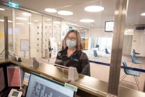 Person wearing mask standing in front of reception desk fitted with glass screen