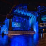 Empty performance stage lit up in blue light. Overhead speakers and acoustic system vaguely visible.