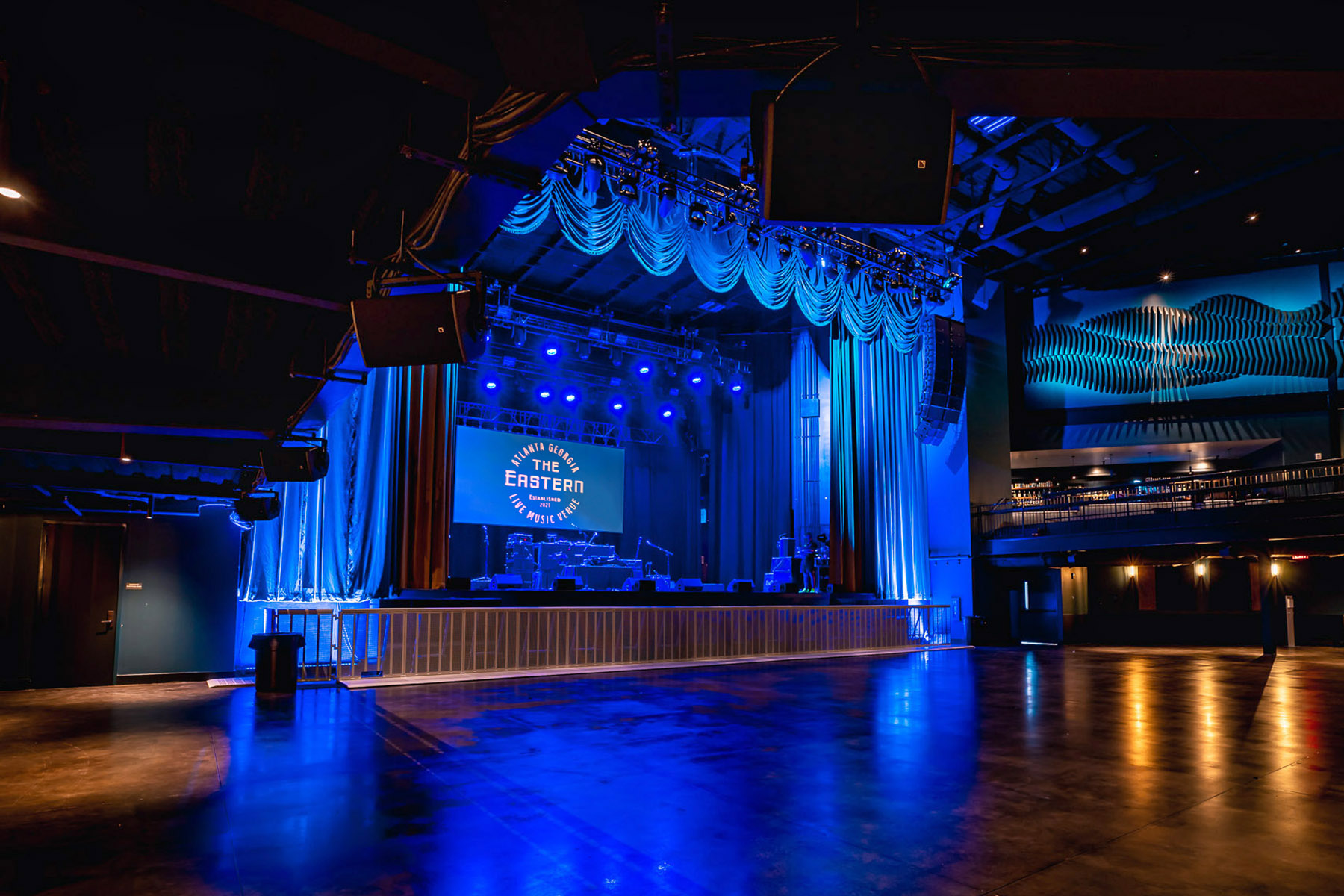 Empty performance stage lit up in blue light. Overhead speakers and acoustic system vaguely visible.