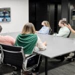 People sitting at a conference table and facing a wall with a screen. The screen has icons of people on a web conferencing platform.