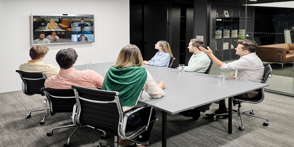 People sitting at a conference table and facing a wall with a screen. The screen has icons of people on a web conferencing platform.