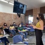 Masked professor addressing a classroom at La Salle Laguna. Above the professor is a screen showcasing remote students