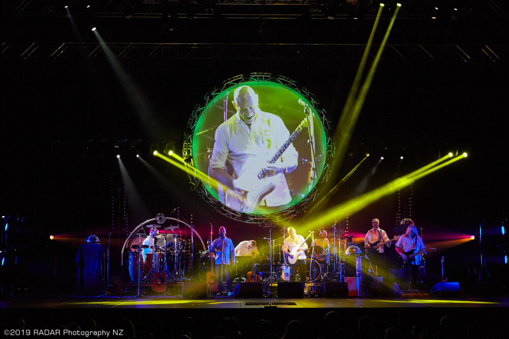 Concert stadium with band playing onstage while giant screen above is showcasing a guitarist.