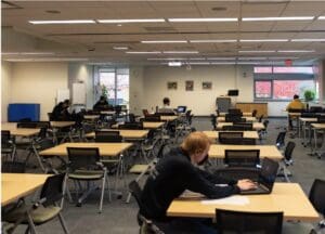 Masked student working on a laptop in a library room.