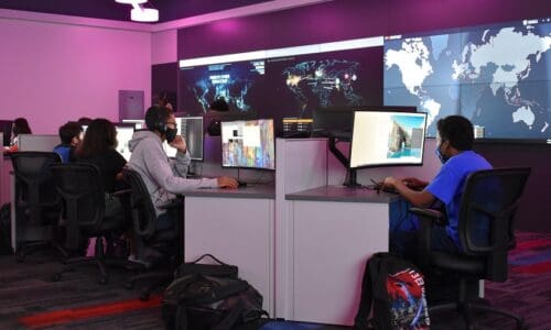 NEISD Cyber Training room with students working on laptops. A video wall with a world map hangs behind powered by RGB Spectrum Galileo processors