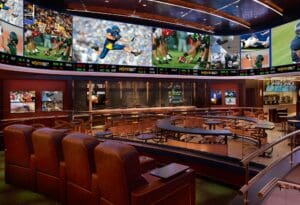 An empty bar with dining seats. Videoscreens are fitted around the perimeter of the bar display different sports along with a sports ticker at the bottom.