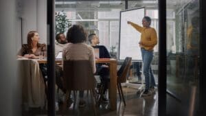 Person presenting on whiteboard to colleagues seated at a table.