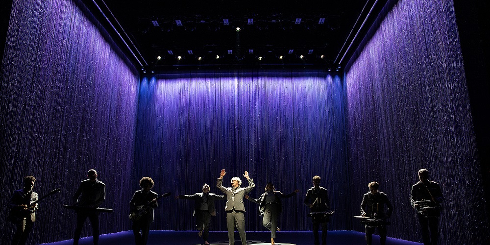 David Byrne standing under the spotlight on American Utopia stage. A line of Meyer Sound LEOPARD loudspeakers are located above him and the orchestra.