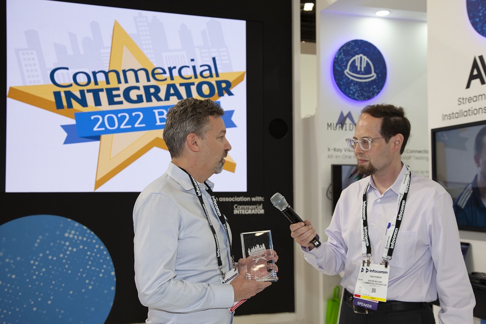 Dan Ferrisi holding mic for Meyer Sound rep at InfoComm. The digital signage behind showcases Commercial Integrator BEST awards logo.