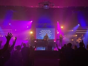 Band performing onstage bathed in purple light. The lyrics of the song appear on a screen behind the singer.