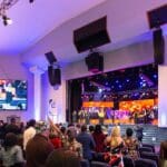 Choir singing on the stage at Celebration Church with loudspeakers hanging on the ceiling.
