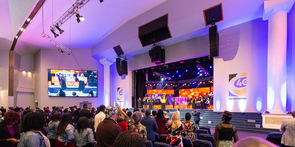 Choir singing on the stage at Celebration Church with loudspeakers hanging on the ceiling.