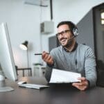 smiling salesperson with headset looking at computer with paper and pen in hand