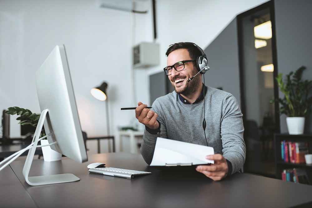 Smiling salesperson with headset looking at computer with paper and pen in hand handling business needs..