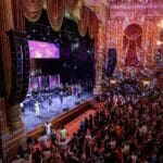 Inside Times Square Church stage where the crowd is performing in front of a packed audience; two loudspeakers are also hanging on either side of the stage.