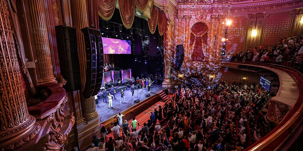 Inside Times Square Church stage where the crowd is performing in front of a packed audience; two loudspeakers are also hanging on either side of the stage.