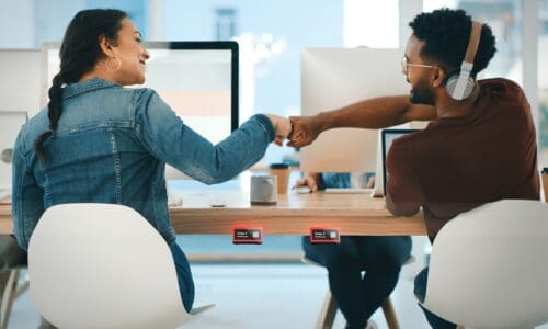 Rearview shot of two young designers giving each other a fist bump in an office, on display is Crestron desk scheduling device