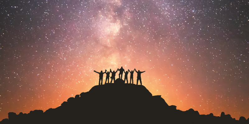 A group of people are standing on the top of the hill next to the Milky Way galaxy holding hands
