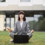 Pretty young female executive practicing yoga, sitting with eyes closed outside office on green grass, in lotus position with hands in mudra gesture, enjoying mental emotional balance during break. sofiko14 / stock.adobe.com