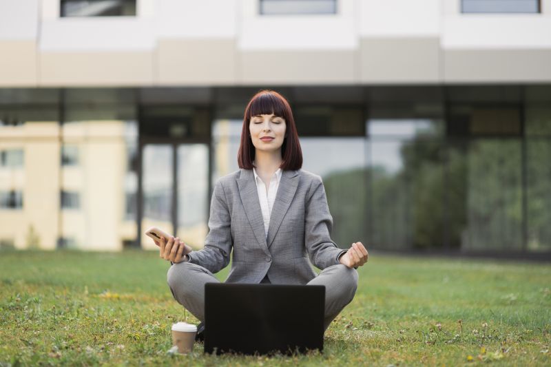 Pretty young female executive practicing yoga, sitting with eyes closed outside office on green grass, in lotus position with hands in mudra gesture, enjoying mental emotional balance during break. sofiko14 / stock.adobe.com