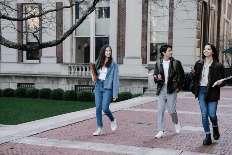 Three college students walking and talking on a campus sidewalk outside a brick university building