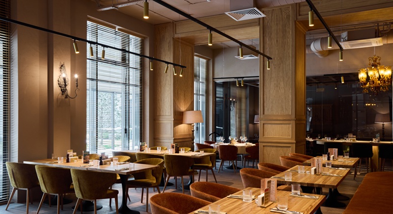 spacious restaurant hall with multiple tables, rowed wooden tables and velvet chairs, tall windows letting